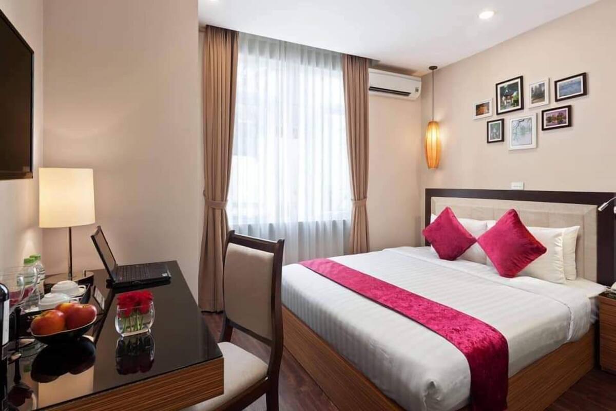 A hotel room with a bed, a desk with a lamp, a chair, a window with curtains, and framed pictures on the wall.