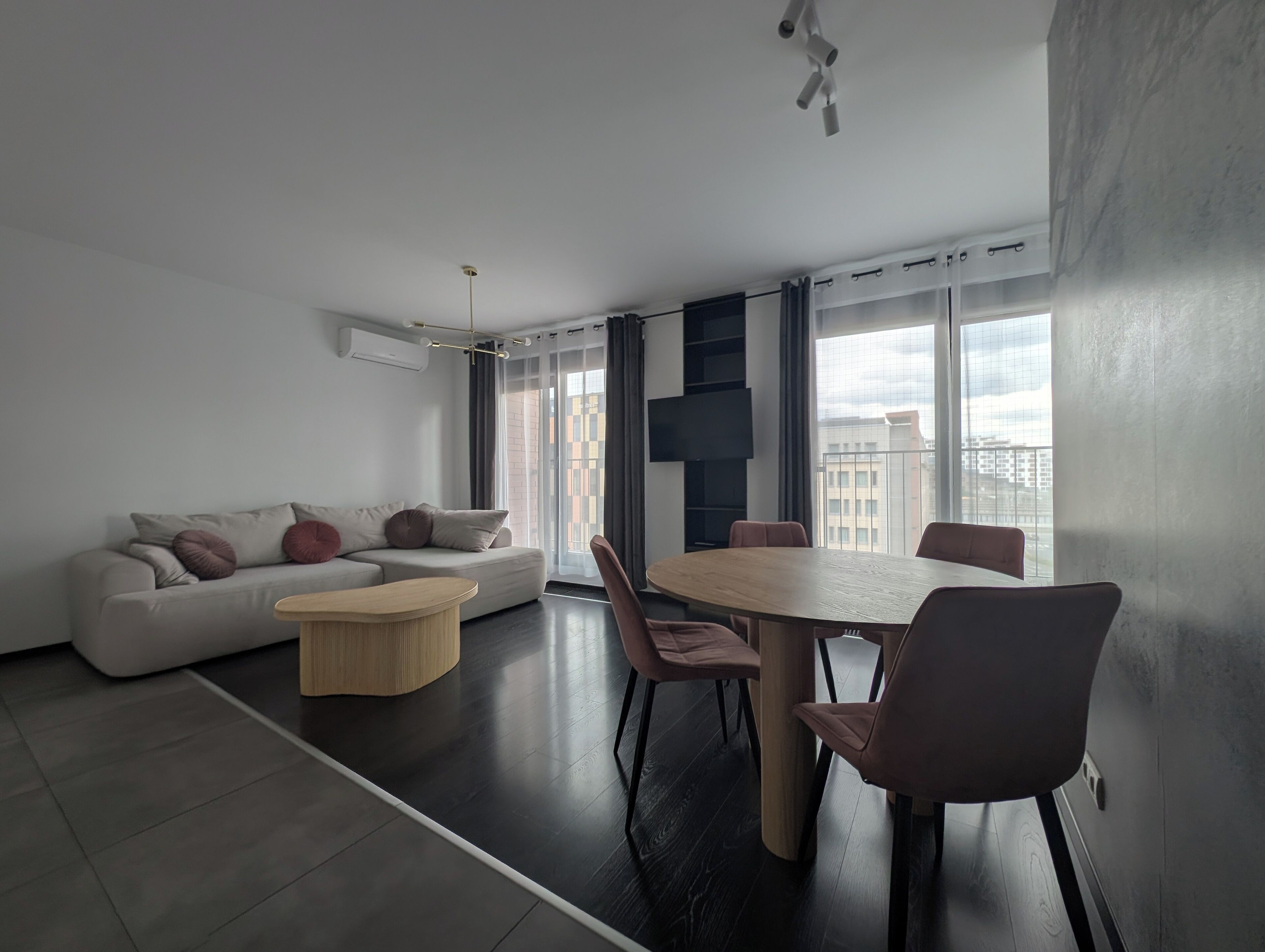 A modern living room with a sofa, a round table, and chairs. There is a large window with a view of buildings.