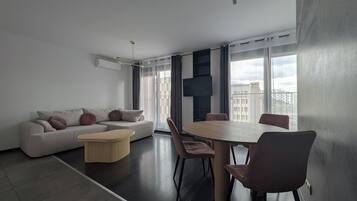 Un soggiorno moderno con un divano, un tavolino rotondo e sedie. C'è un'ampia finestra che si affaccia su degli edifici.