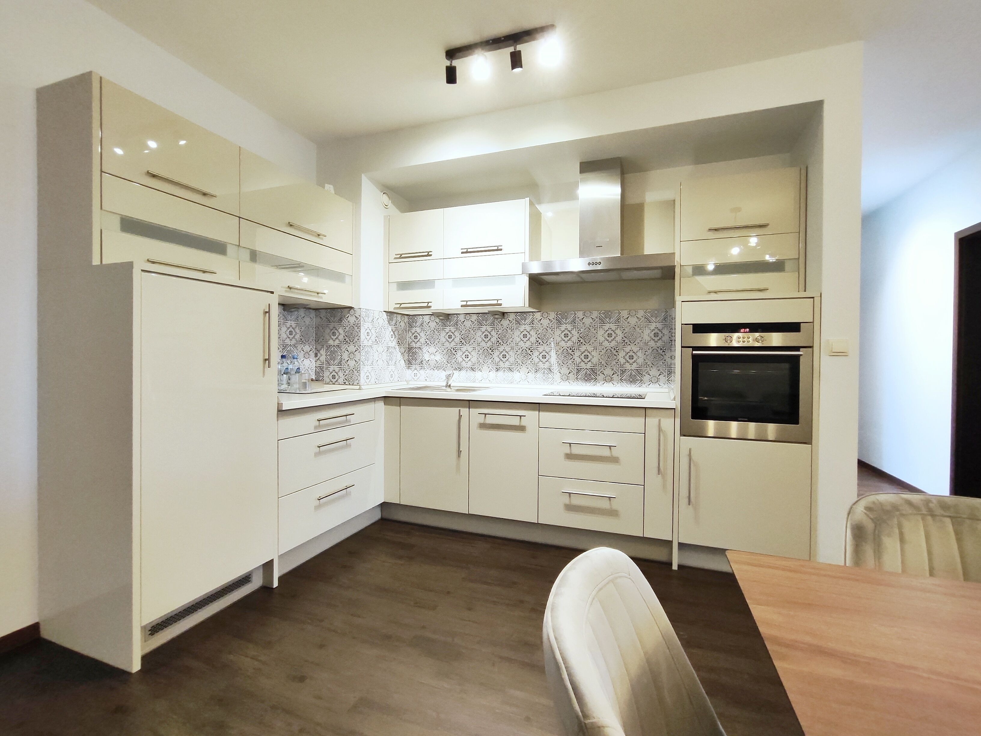 Une cuisine moderne avec des meubles blancs, un four encastré et un coin repas comprenant une table en bois et une chaise.