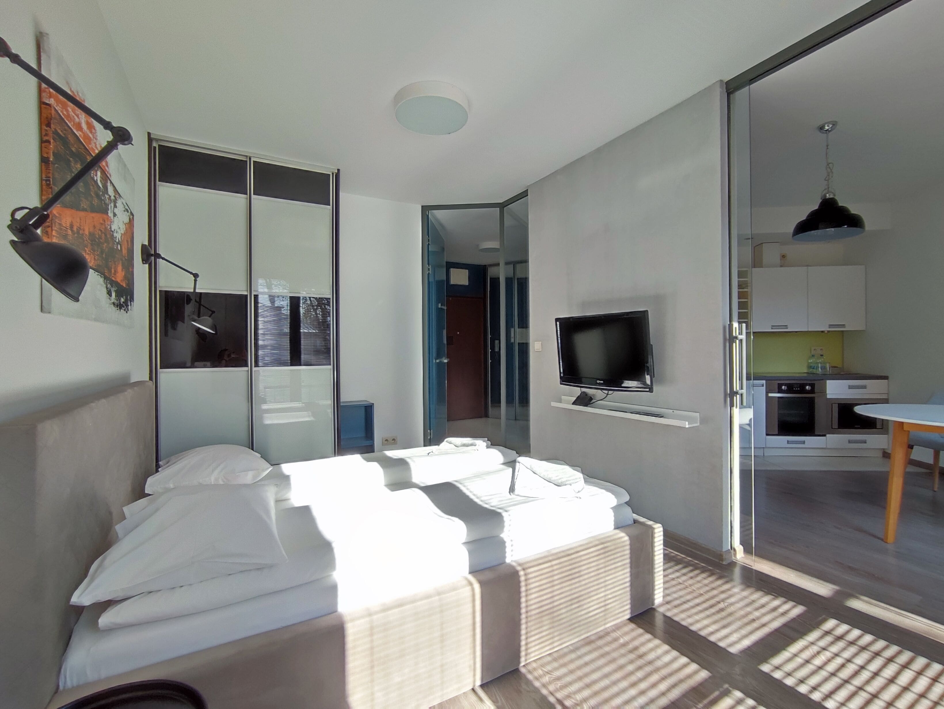 A modern bedroom with a bed, a TV, a small kitchenette, and a sliding glass door leading to a bathroom.