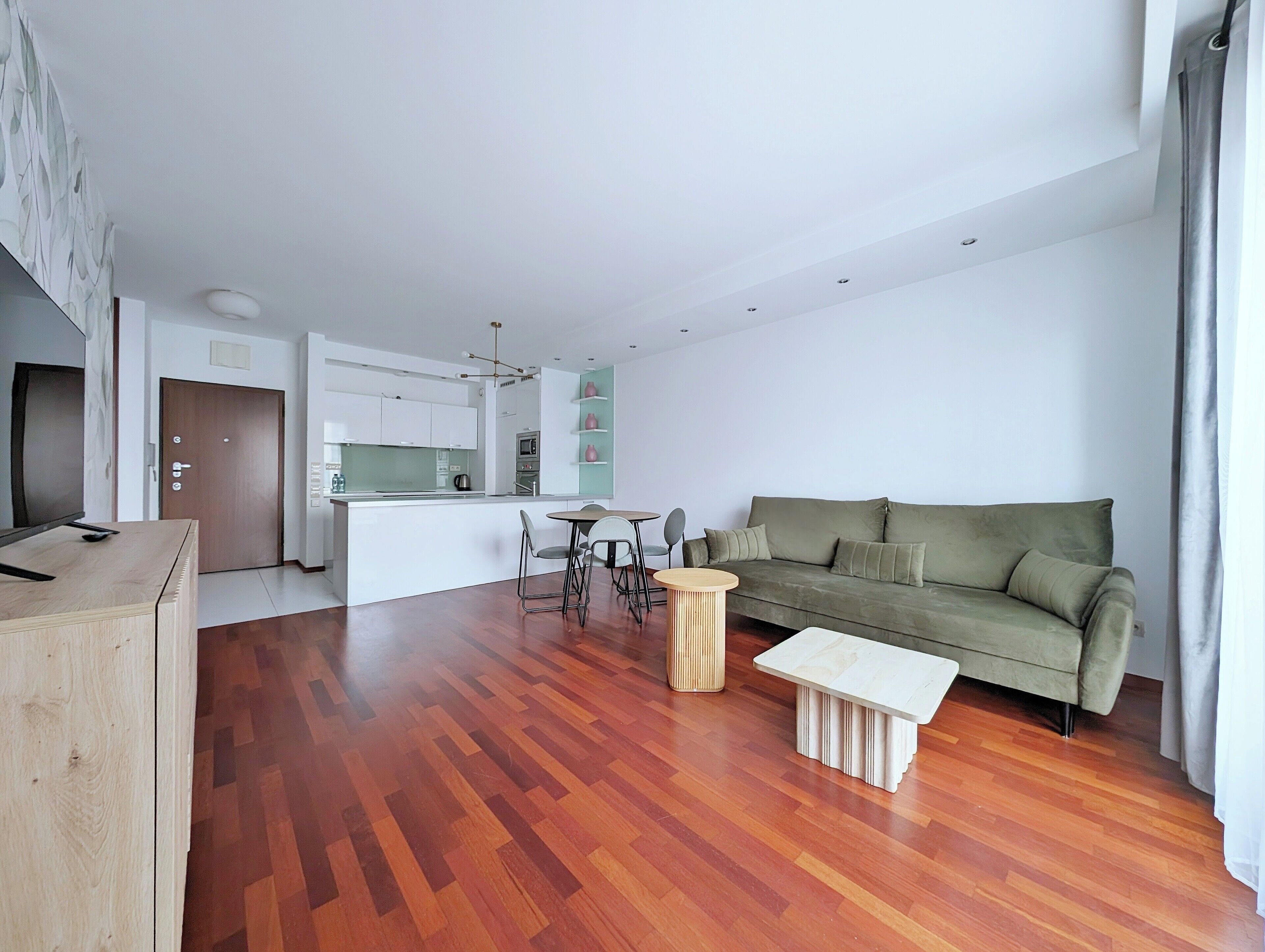 Een moderne woonkamer met houten vloer, een groene bank, een witte salontafel en een keuken met witte kasten en een eettafel.
