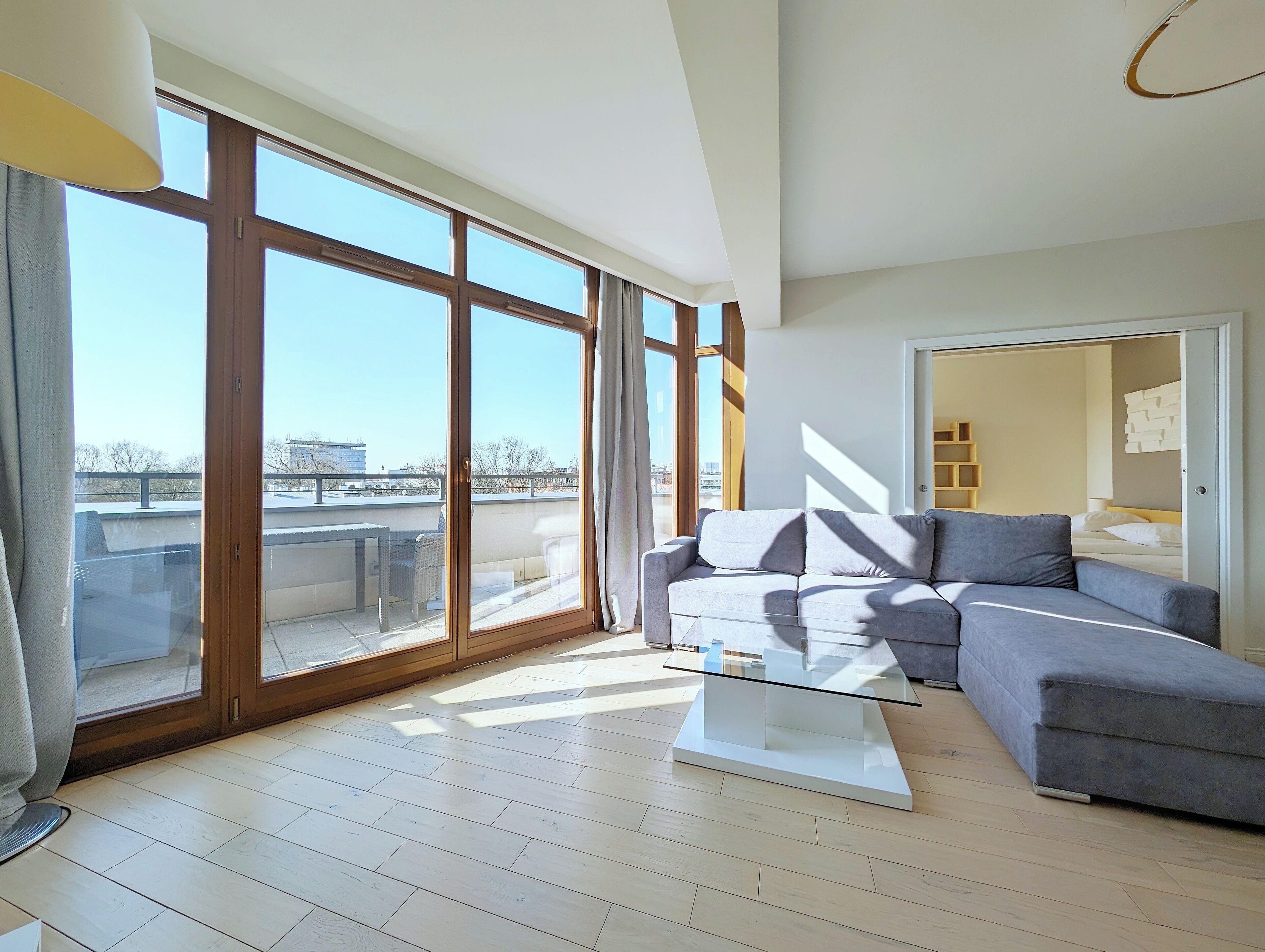 A spacious living room with a large sliding glass door leading to a balcony, a grey sofa, a glass coffee table, and a modern interior design.