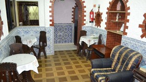 Una habitación con piso a cuadros, paredes enlosadas, un panel mural azul y blanco, un sillón a cuadros, una pequeña mesa con mantel blanco y una silla de madera.