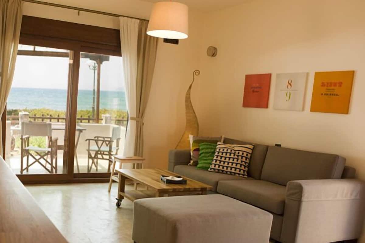 Uma sala de estar moderna com sofá, banqueta e mesa de centro. Há uma porta de vidro deslizante que dá para uma área externa com vista para o mar.