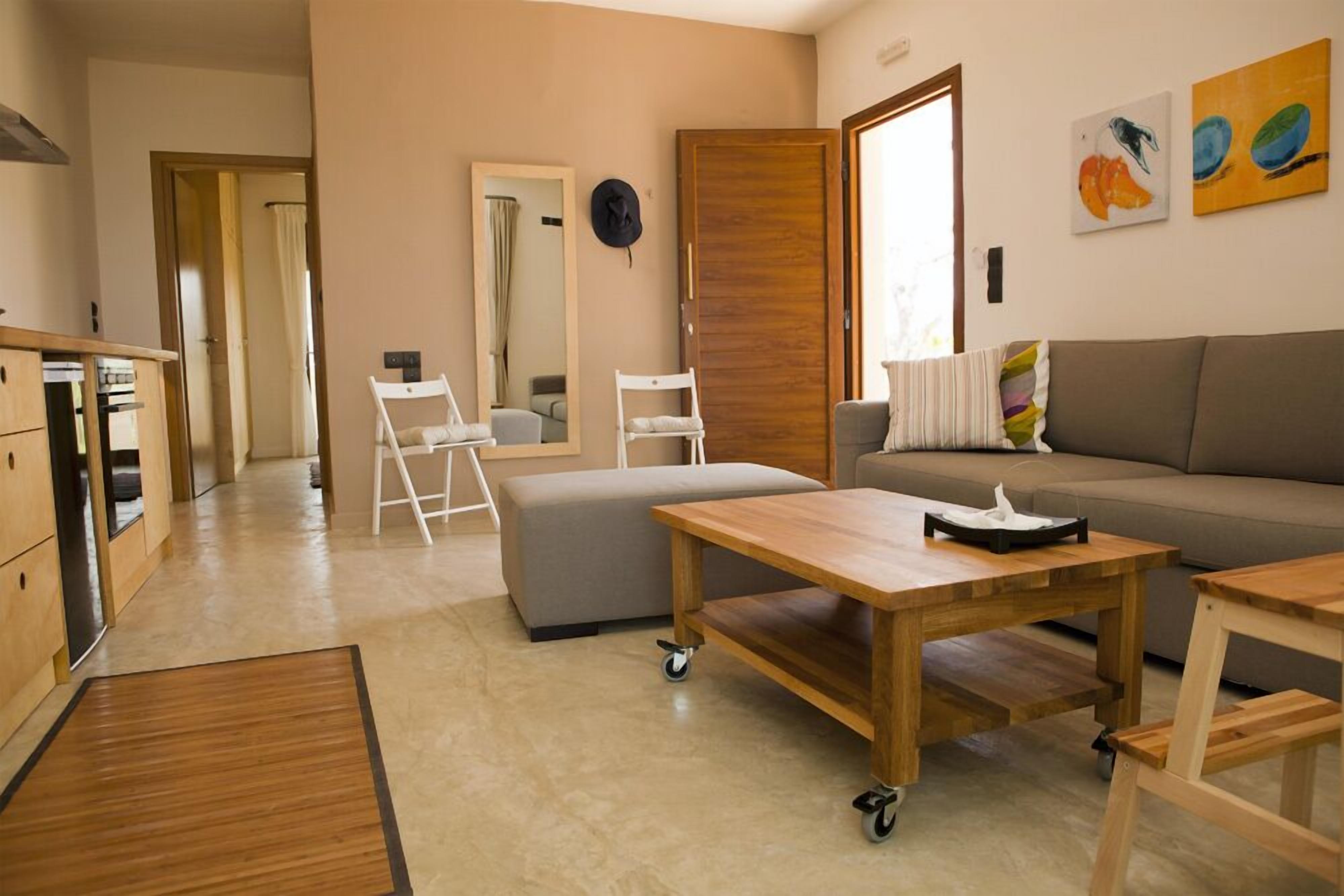 A modern living room with a sofa, a wooden coffee table, a small ottoman, and a kitchen area with cabinets and a countertop.