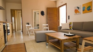 Uma sala de estar moderna com sofá, mesa de centro de madeira, um pequeno banquinho e uma área de cozinha com armários e bancada.
