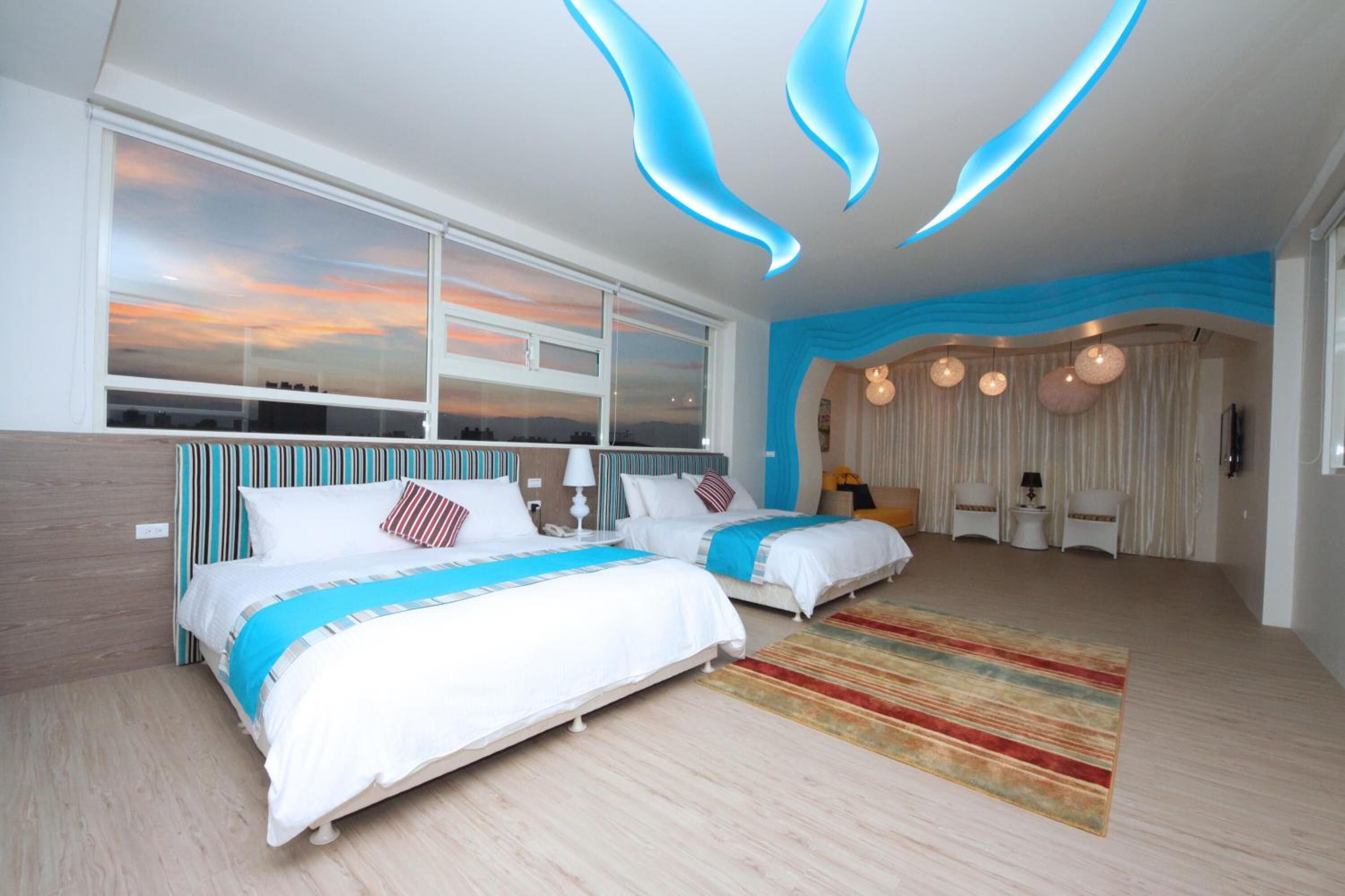 Una habitación de hotel moderna con dos camas, un gran ventanal con vistas a la ciudad, una pared de acento azul y una zona de estar acogedora con una lámpara y una mesita.