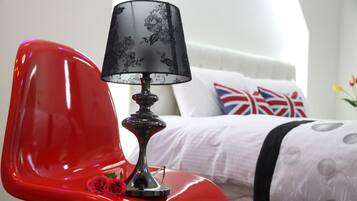 Uma cadeira vermelha com uma luminária preta ao lado, uma cama branca com almofada da Union Jack e uma parede branca.