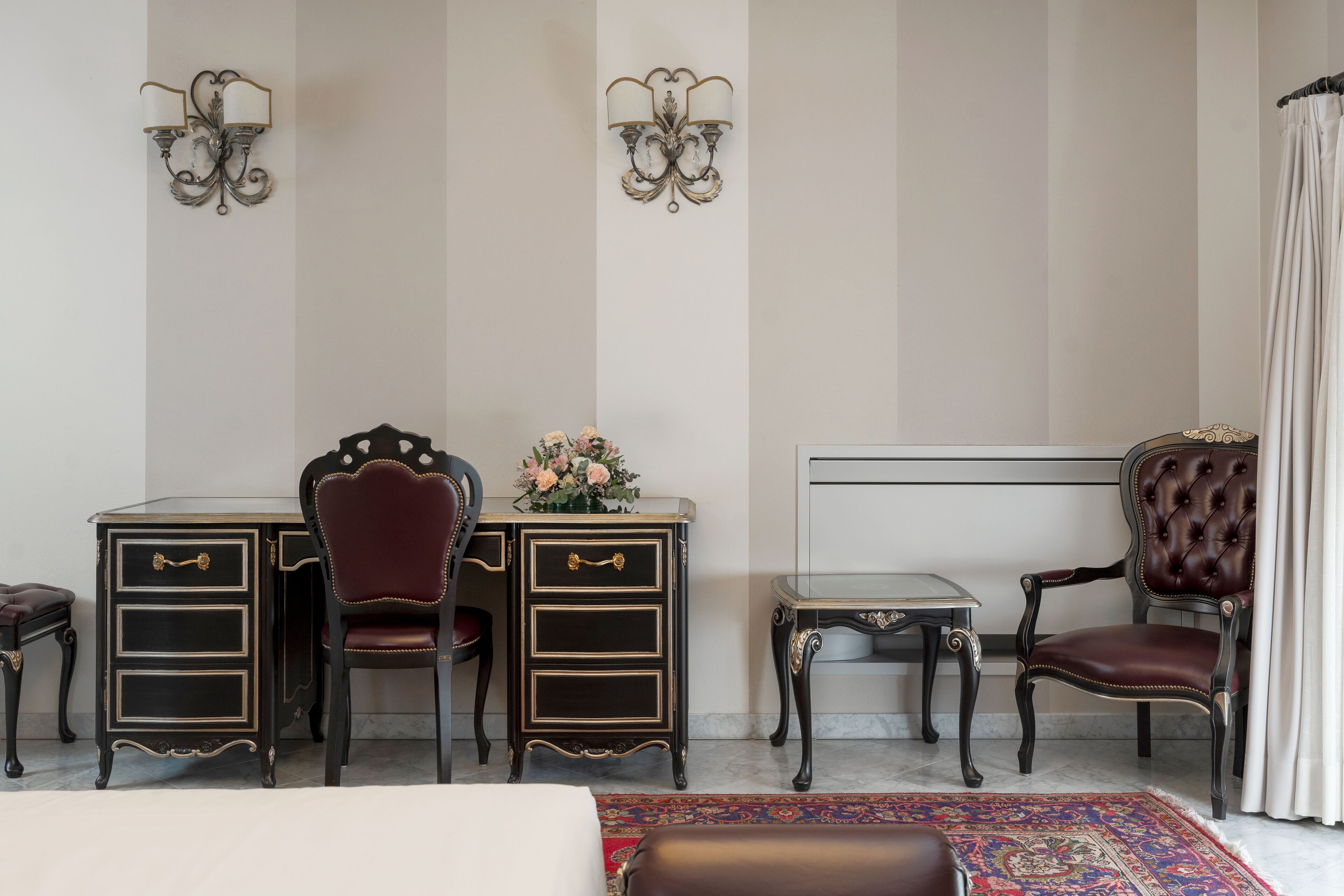 A room with a desk, two chairs, a small table, and a decorative vase with flowers.