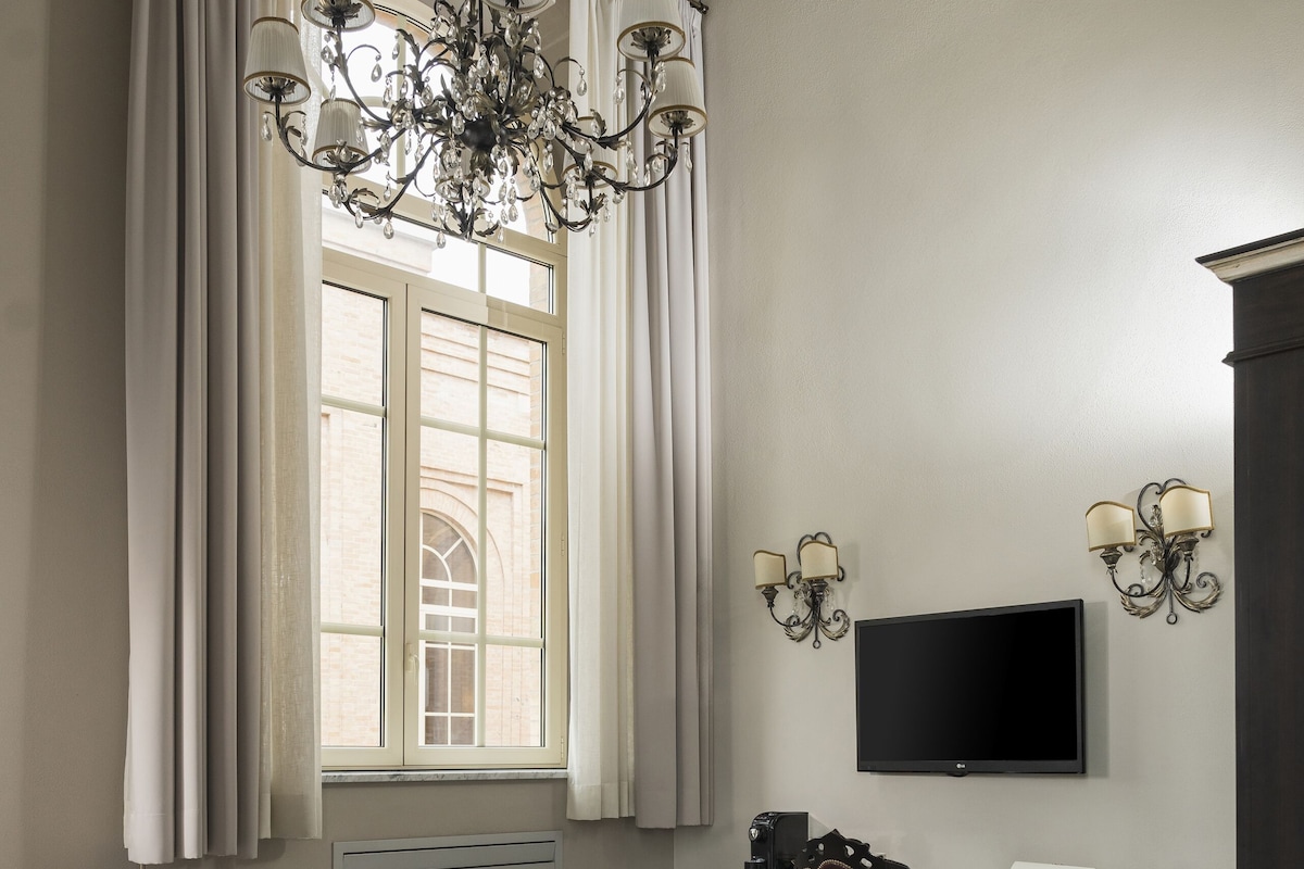 A hotel room with a large bed, a desk with a chair, a television, and a chandelier.