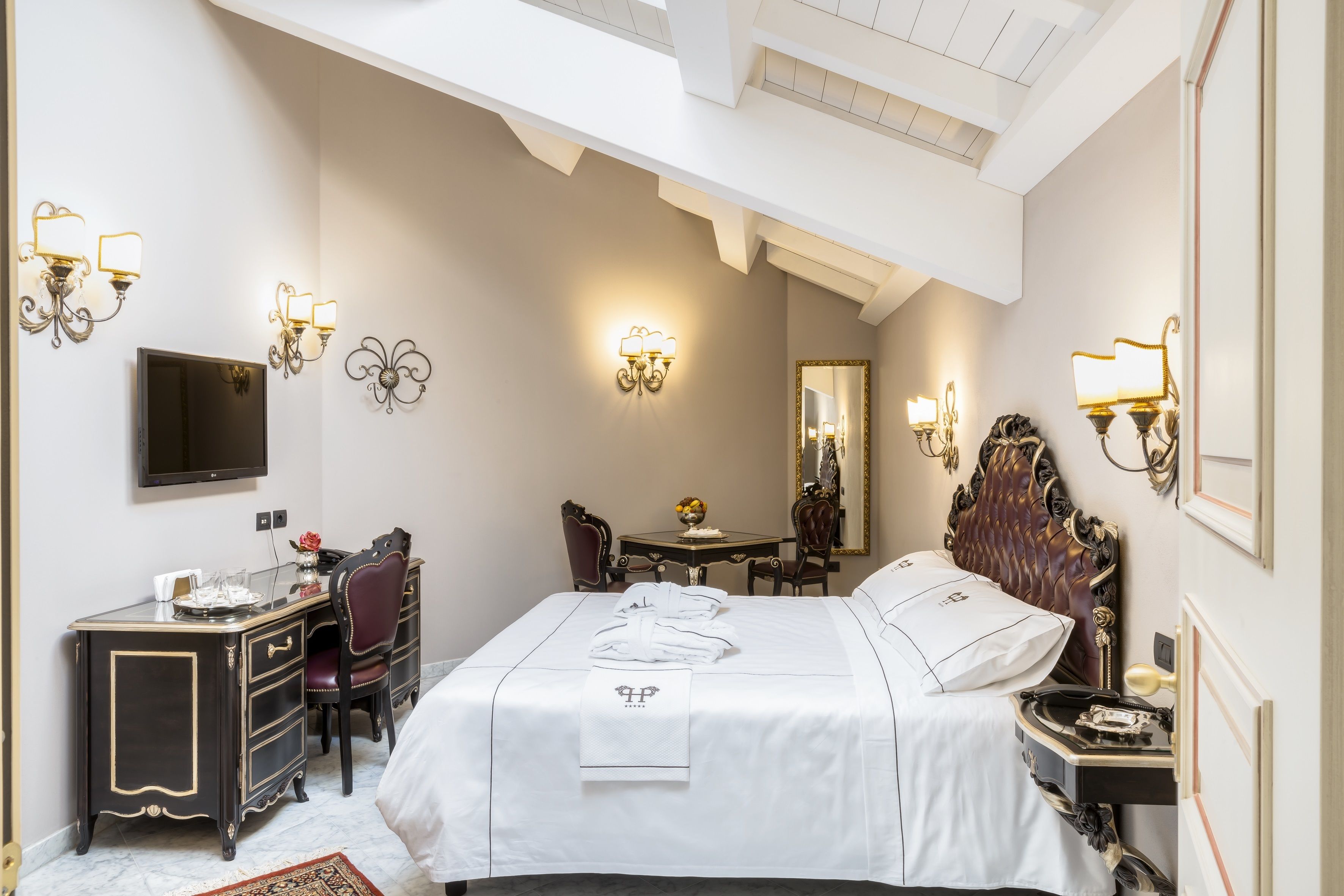 A hotel room with a bed, a desk, a chair, a TV, and wall sconces.