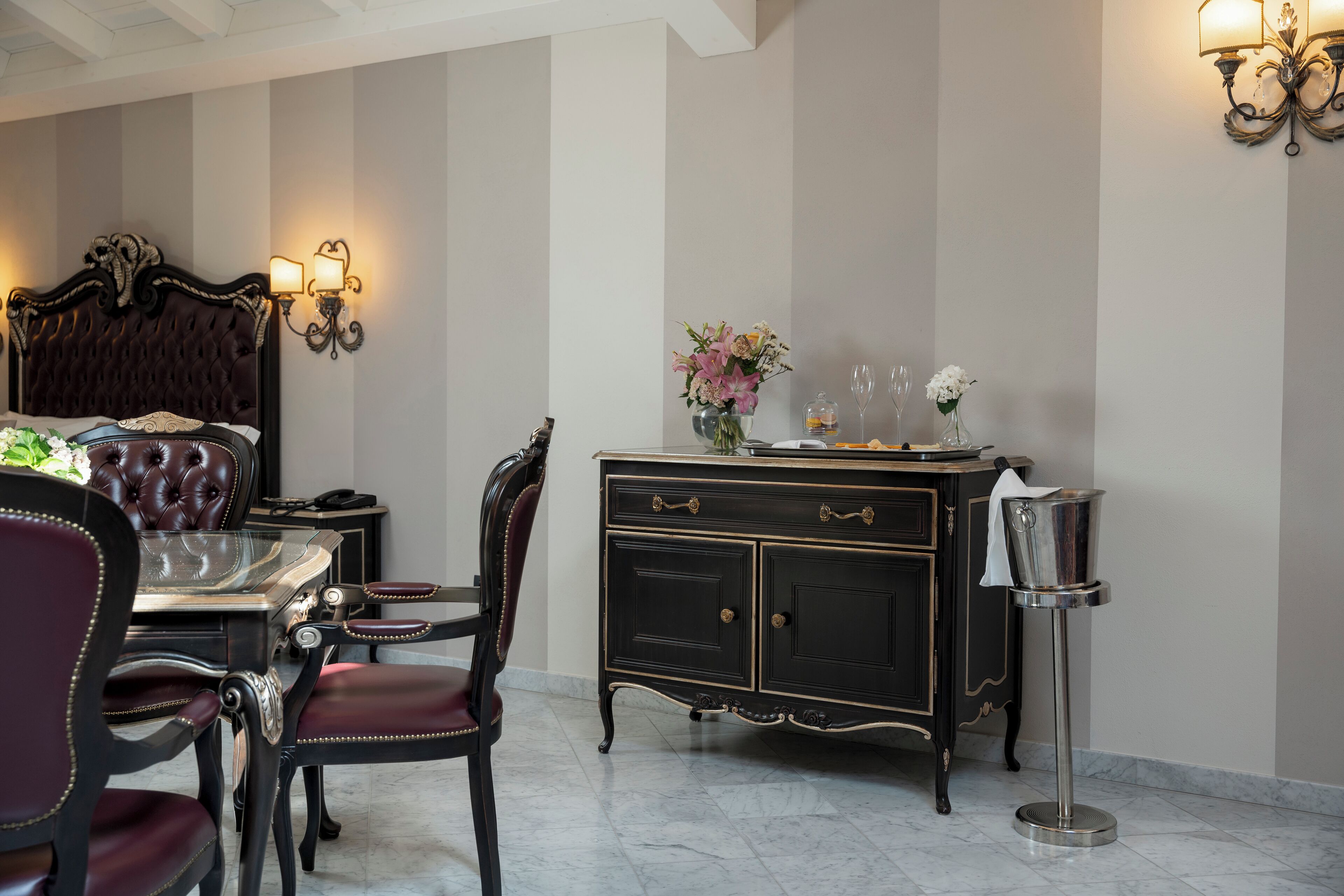 A room with a bed, a dining table with chairs, a side table with a vase of flowers, and a wall-mounted lamp.