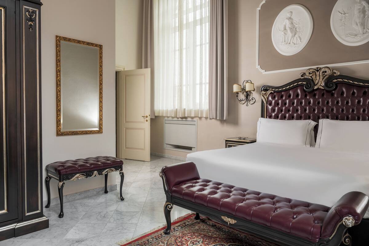 Une chambre à coucher avec un grand lit, un banc, une armoire, un miroir et une porte.