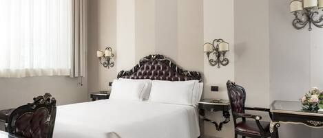 A hotel room with a large bed, a desk with a chair, a chandelier, and wall sconces.