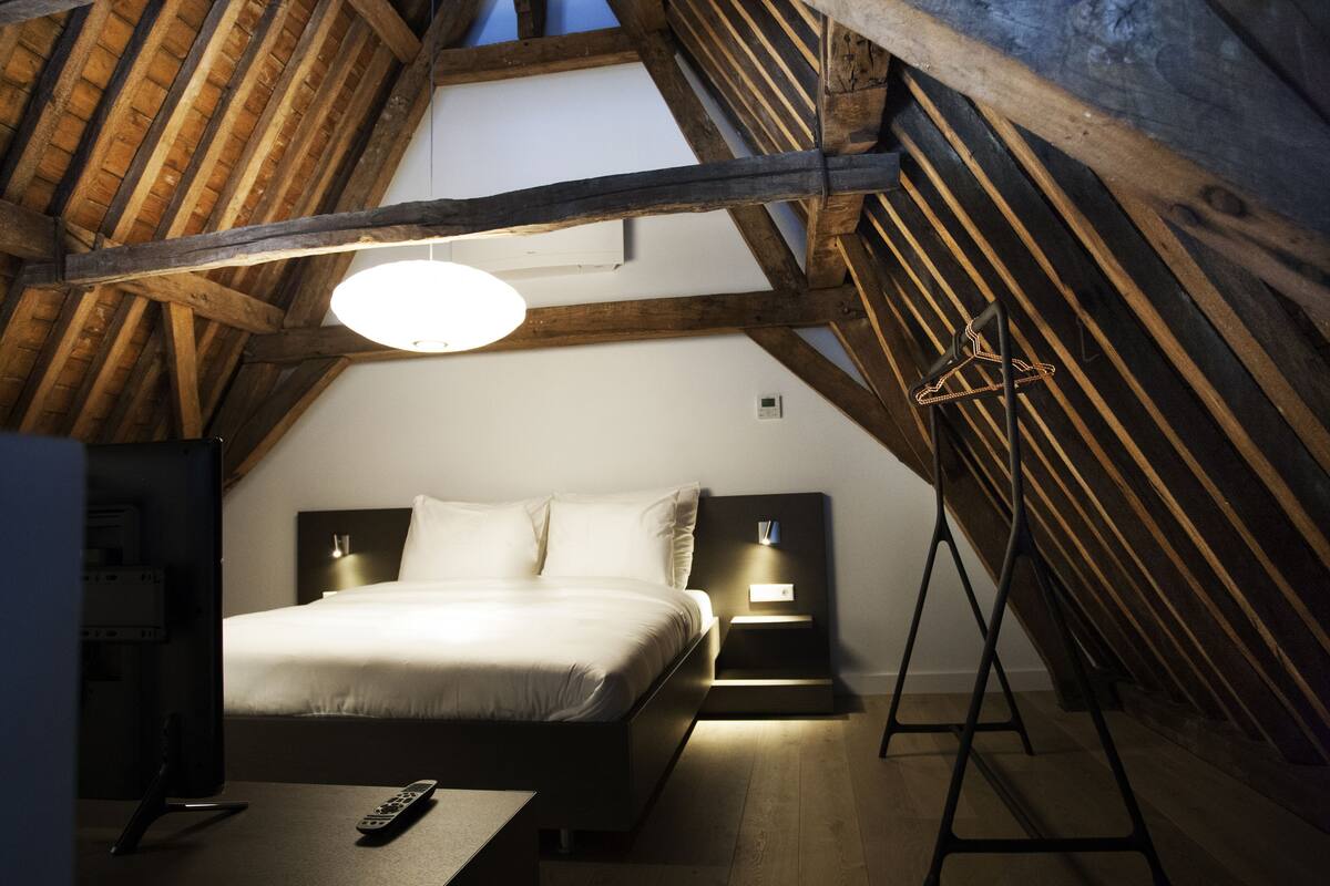 A bedroom with a wooden ceiling, a bed with white bedding, bedside lamps, and a remote control on the floor.