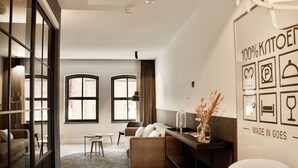 A modern living room with a sofa, a coffee table, and a glass partition. There are two windows with curtains, a wall with a '100% KNOXED' sign, and a 'MADE IN GOES' sign. - Hotel & Brasserie Katoen (Goes)