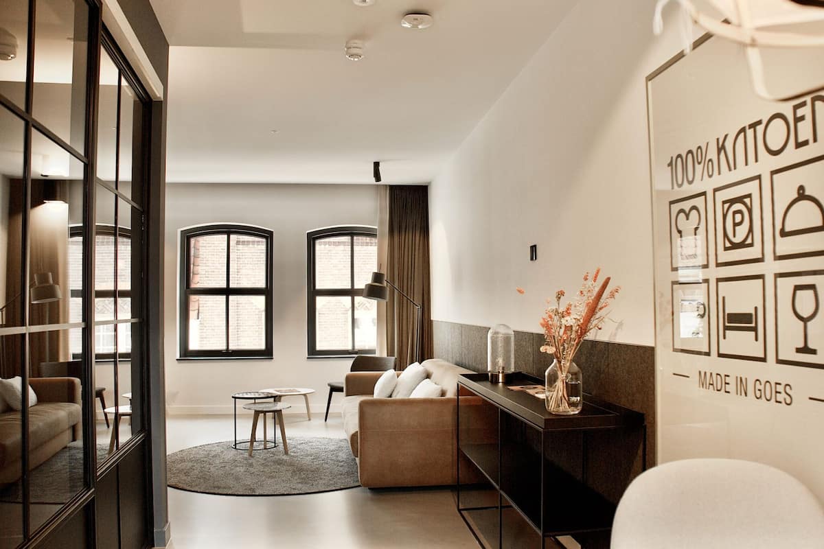 A modern living room with a sofa, a coffee table, and a glass partition. There are two windows with curtains, a wall with a '100% KNOXED' sign, and a 'MADE IN GOES' sign.