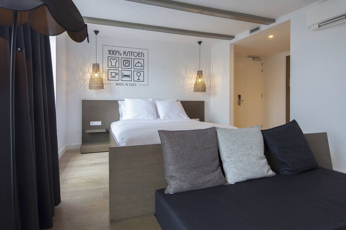 A modern hotel room with a neatly made bed, a sofa, and a wall with a '100% KANTOEN MADE IN GOES' sign.