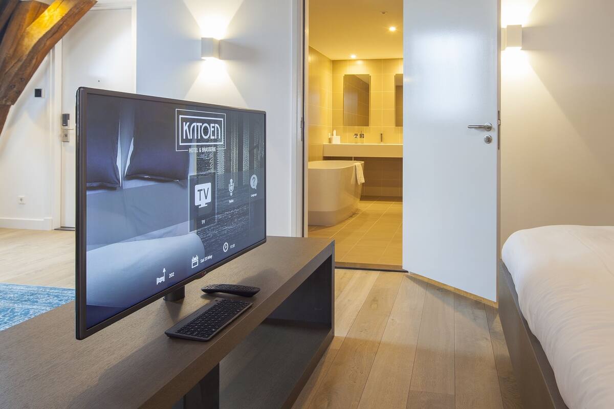 A modern hotel room with a TV, wooden flooring, a bed, and a wall sign indicating '100% KATOEN MADE IN GOES'.