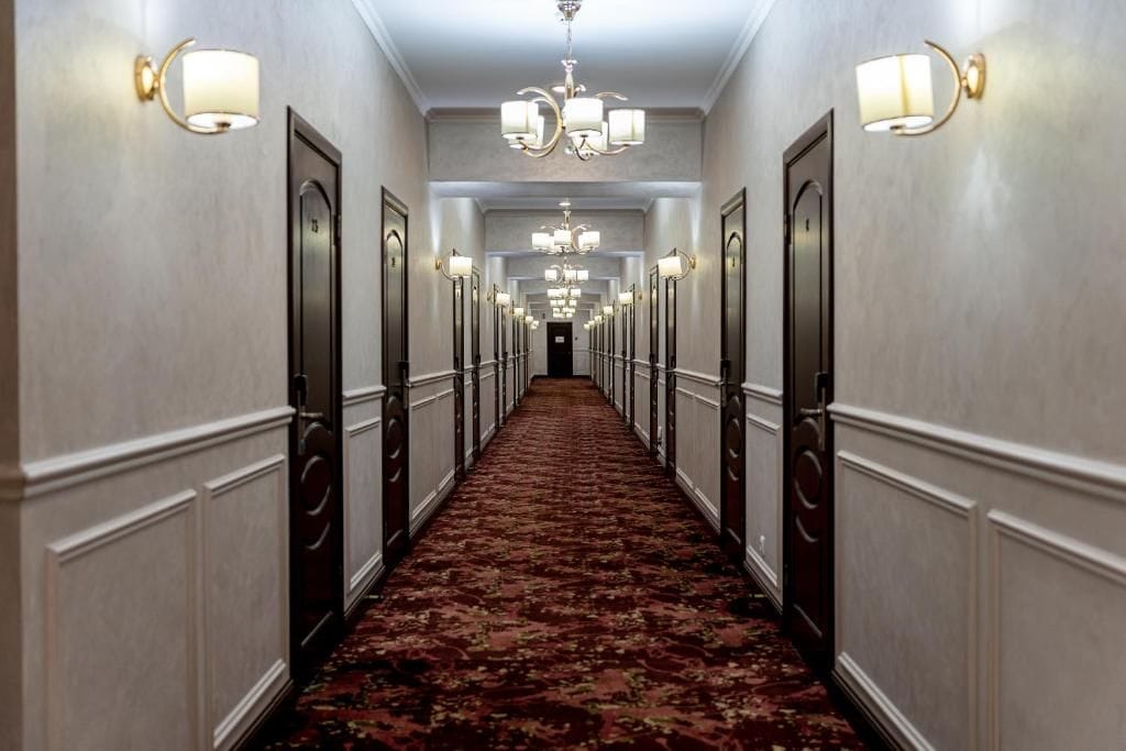 A hotel hallway with a carpeted floor, doors on both sides, and wall-mounted light fixtures.