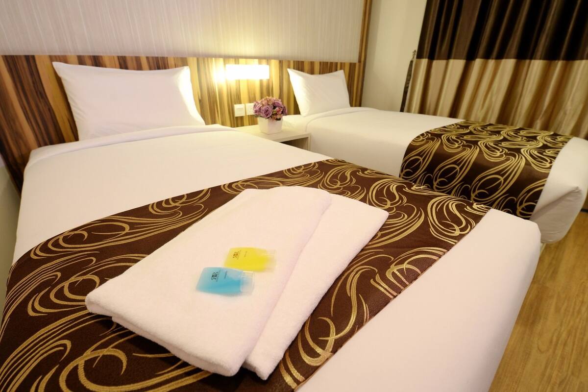 Une chambre d’hôtel avec deux lits, chacun recouvert d’une couvre-lit marron à motifs et agrémenté de coussins blancs. On y trouve une table de chevet avec une lampe et un vase de fleurs.