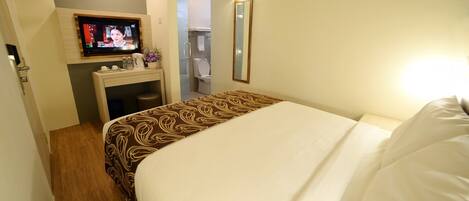 Habitación de hotel con cama, televisor montado en la pared, escritorio con espejo y baño visible a través de una puerta abierta.