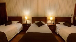Hotellihuone, jossa on kolme erillistä sänkyä. Jokaisessa sängyssä on puinen pääty, yöpöytä ja lamppu.
