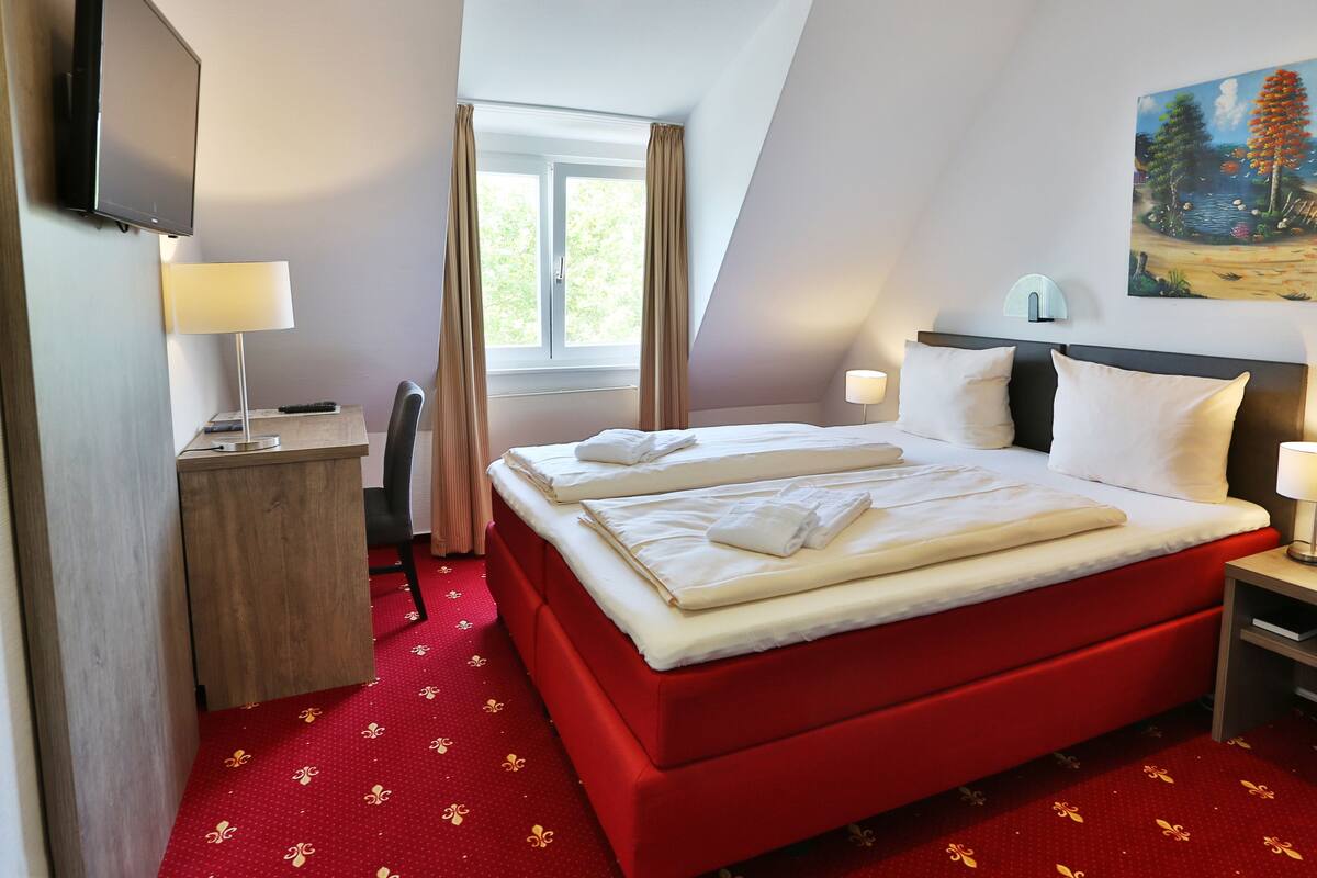 A hotel room with a red bed, white linens, a desk with a lamp, a chair, and a painting on the wall.