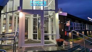 Moderni hotellin sisäänkäynti, jossa on lasijulkisivu, neonvalomainos ja portaat, jotka johtavat ovelle.
