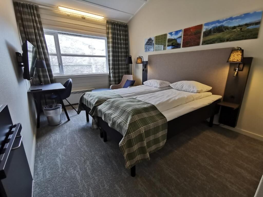 A hotel room with a bed, a desk, a chair, a lamp, and a window with curtains.