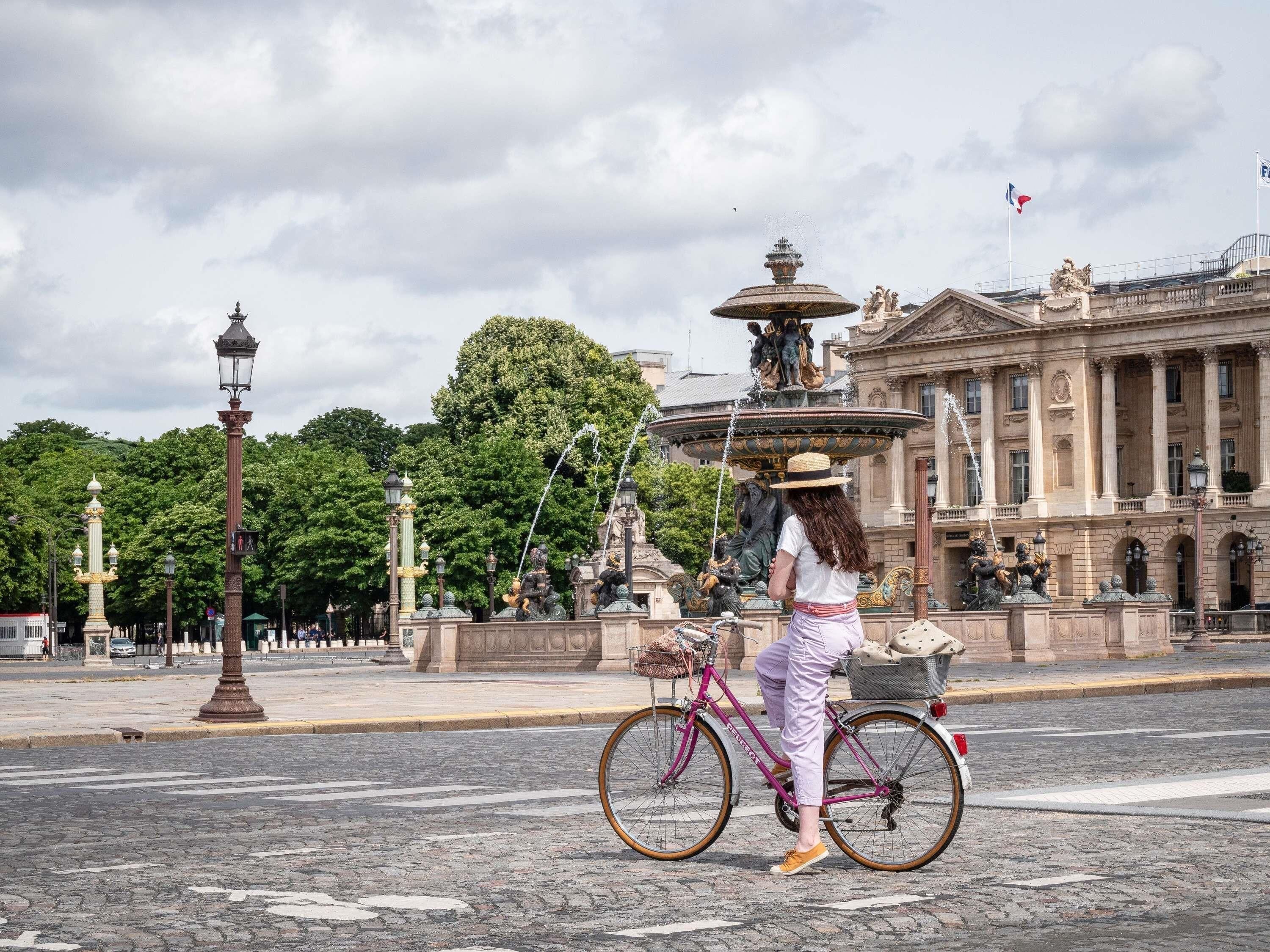 A person on a bicycle with a basket, a fountain, and a building with columns in the background.