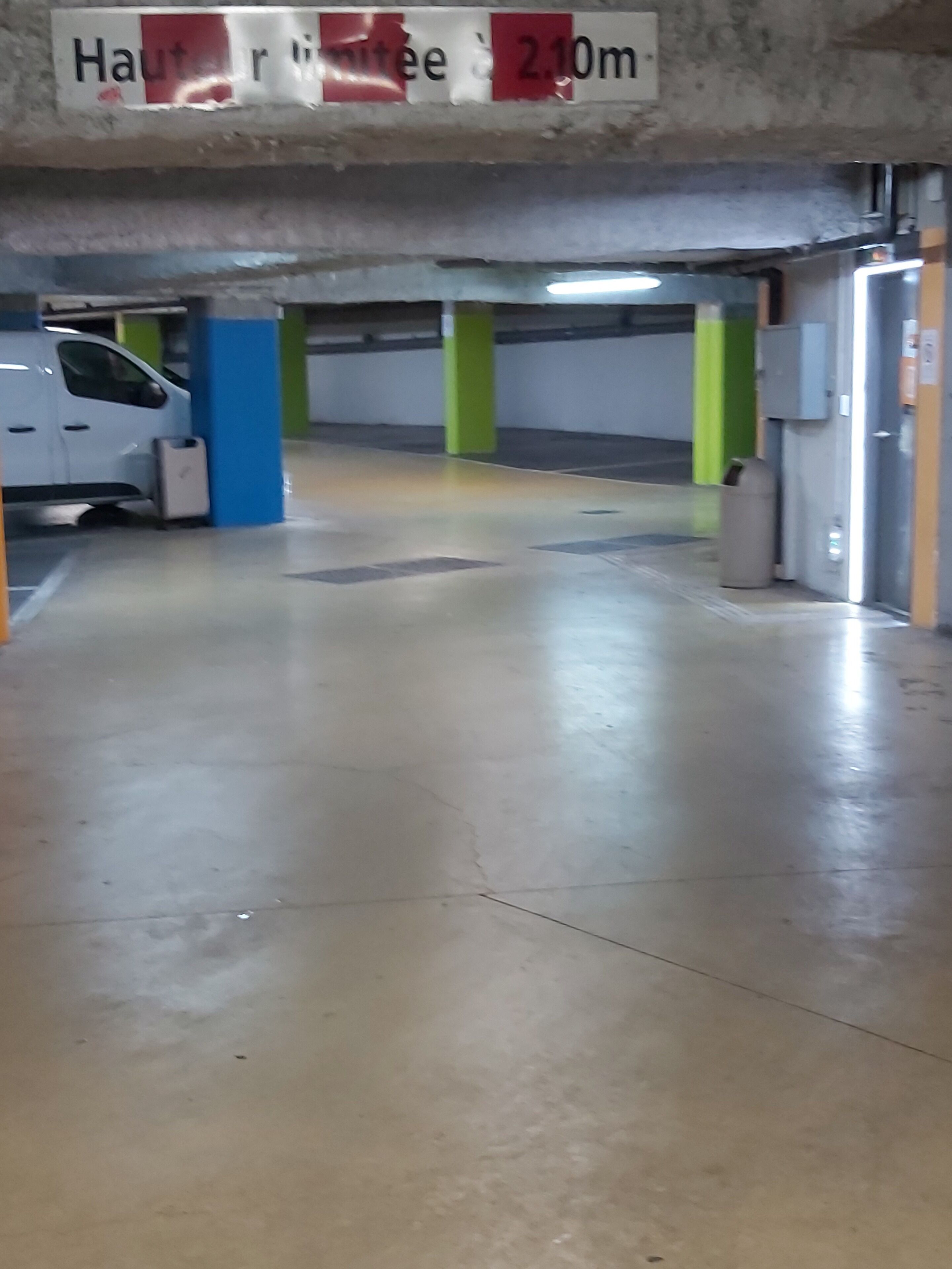 A parking garage with a white van parked near a blue pillar, a green pillar, and a trash bin.
