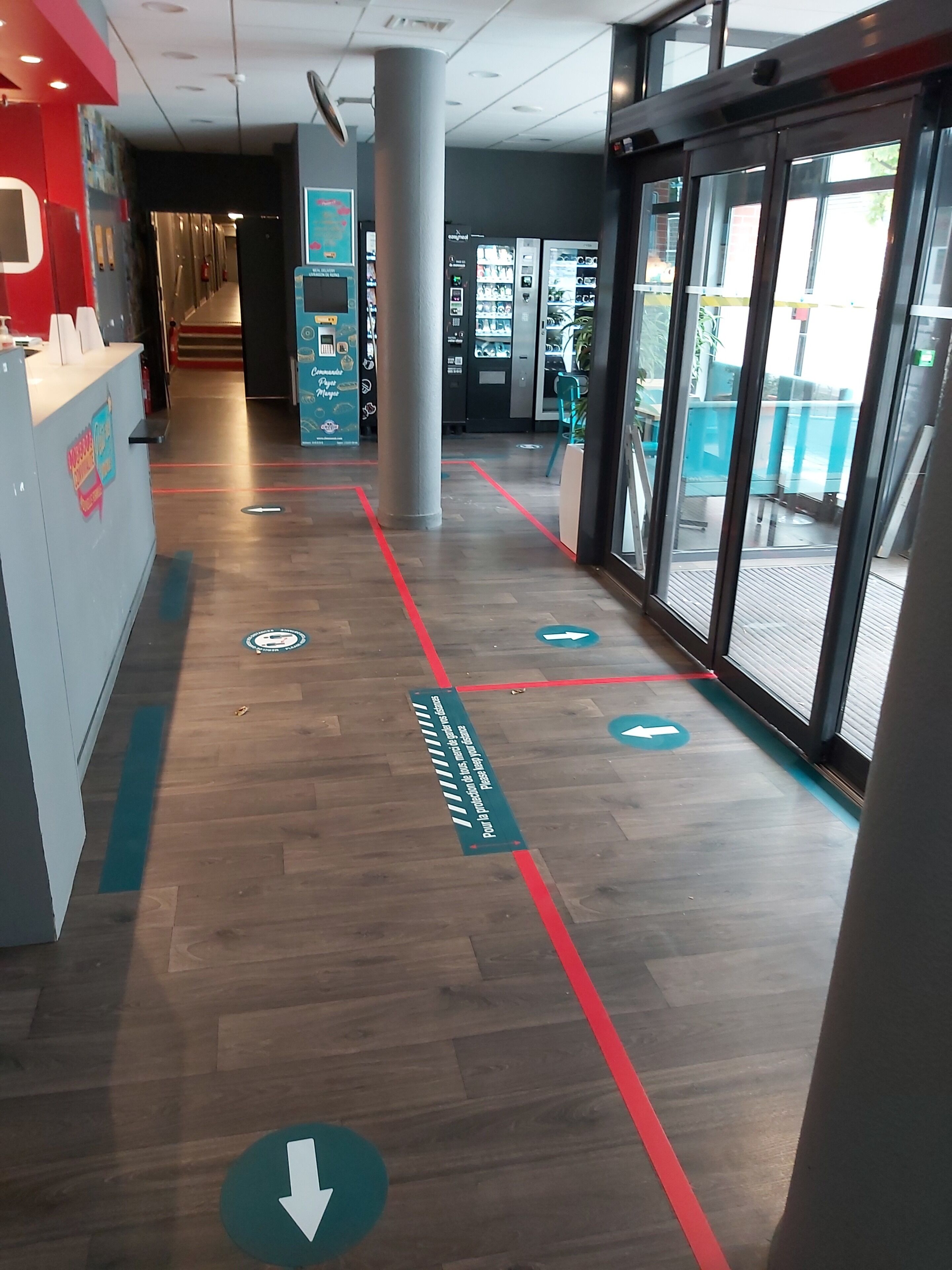 Hotelgang med gulvpile, reception, automater og en glasdør til udendørs.