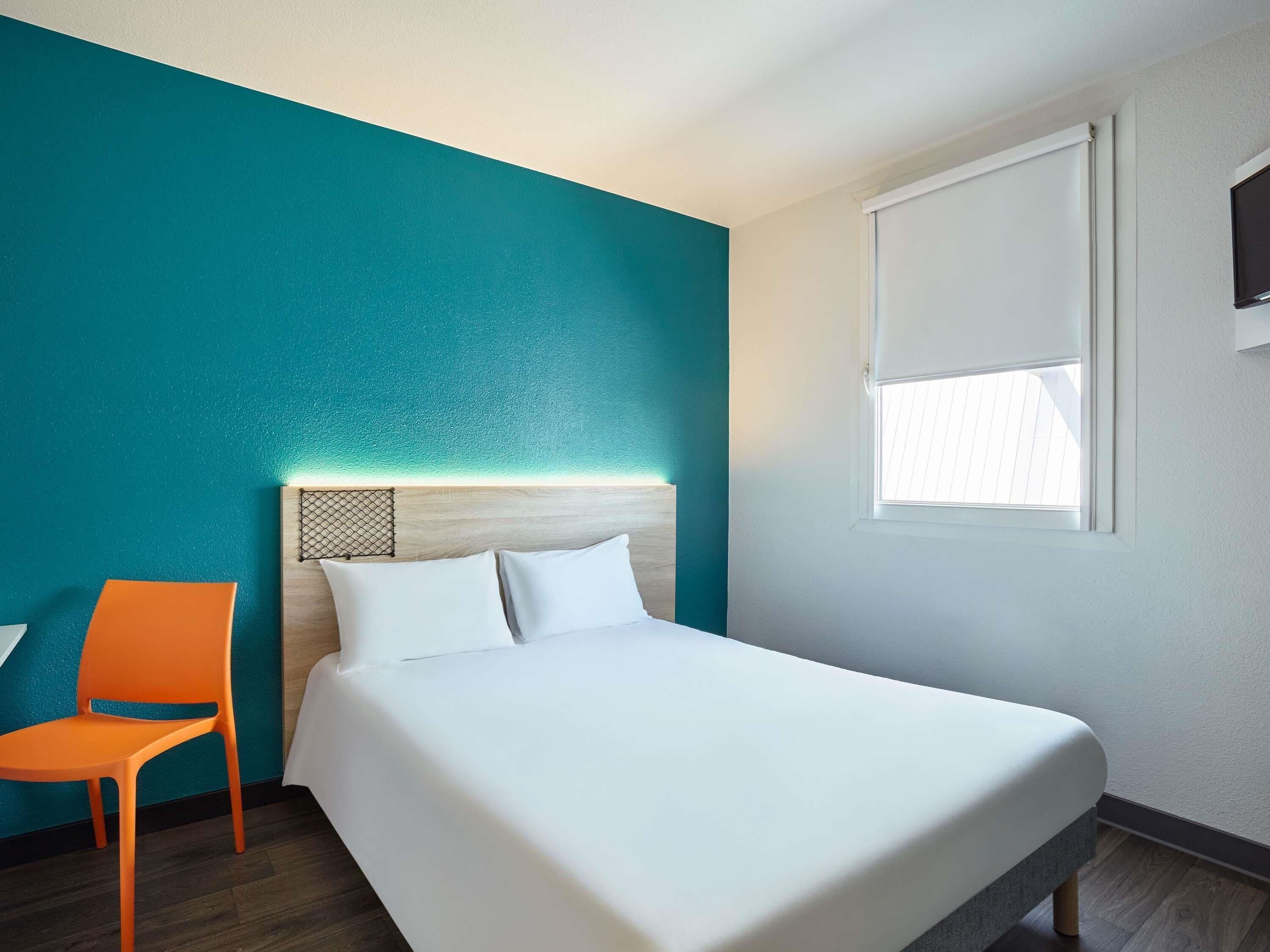 A hotel room with a bed, a chair, a window, and teal walls.