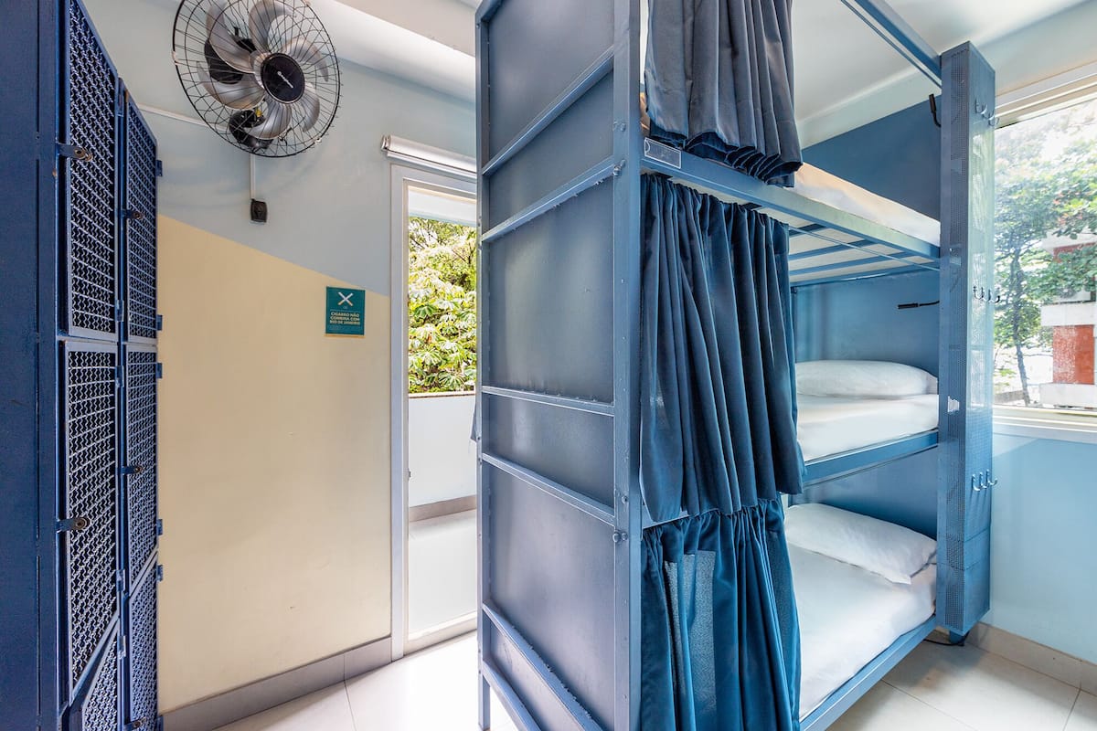 Quarto com beliche, um ventilador, uma porta com placa e vista para o verde lá fora.