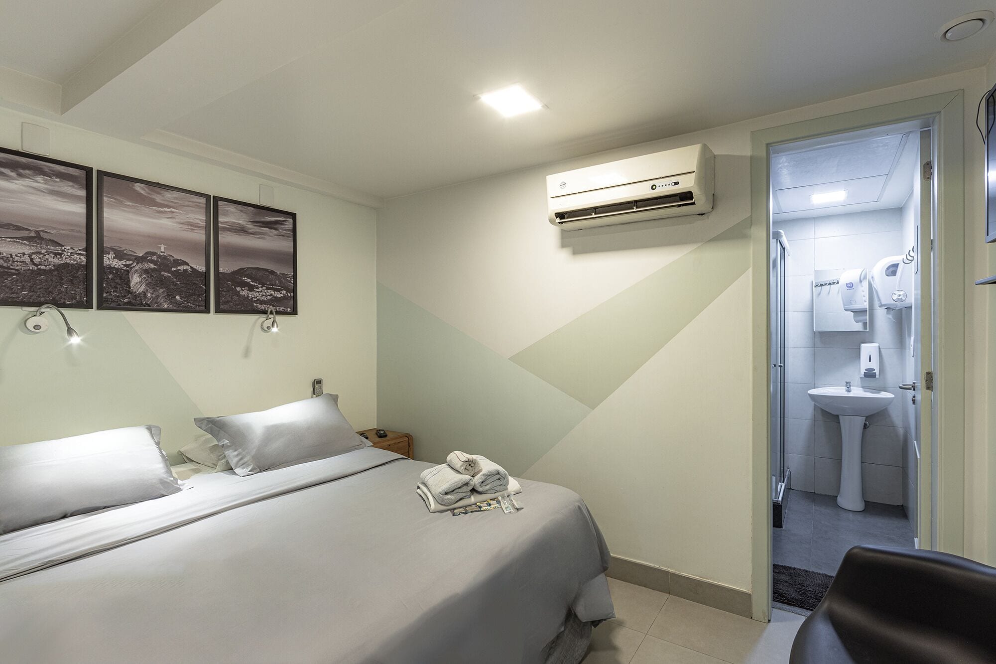 Habitación de hotel con cama, aire acondicionado de pared, baño con lavamanos y espejo, y una puerta que da a otra habitación.