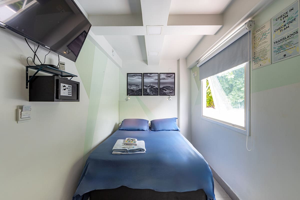 Quarto pequeno com cama, televisão, telefone e janela com vista para área verde.