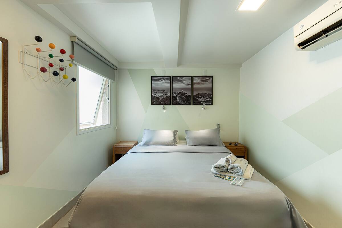 Quarto de hotel com cama, duas mesas de cabeceira, uma janela e ar condicionado fixado na parede.