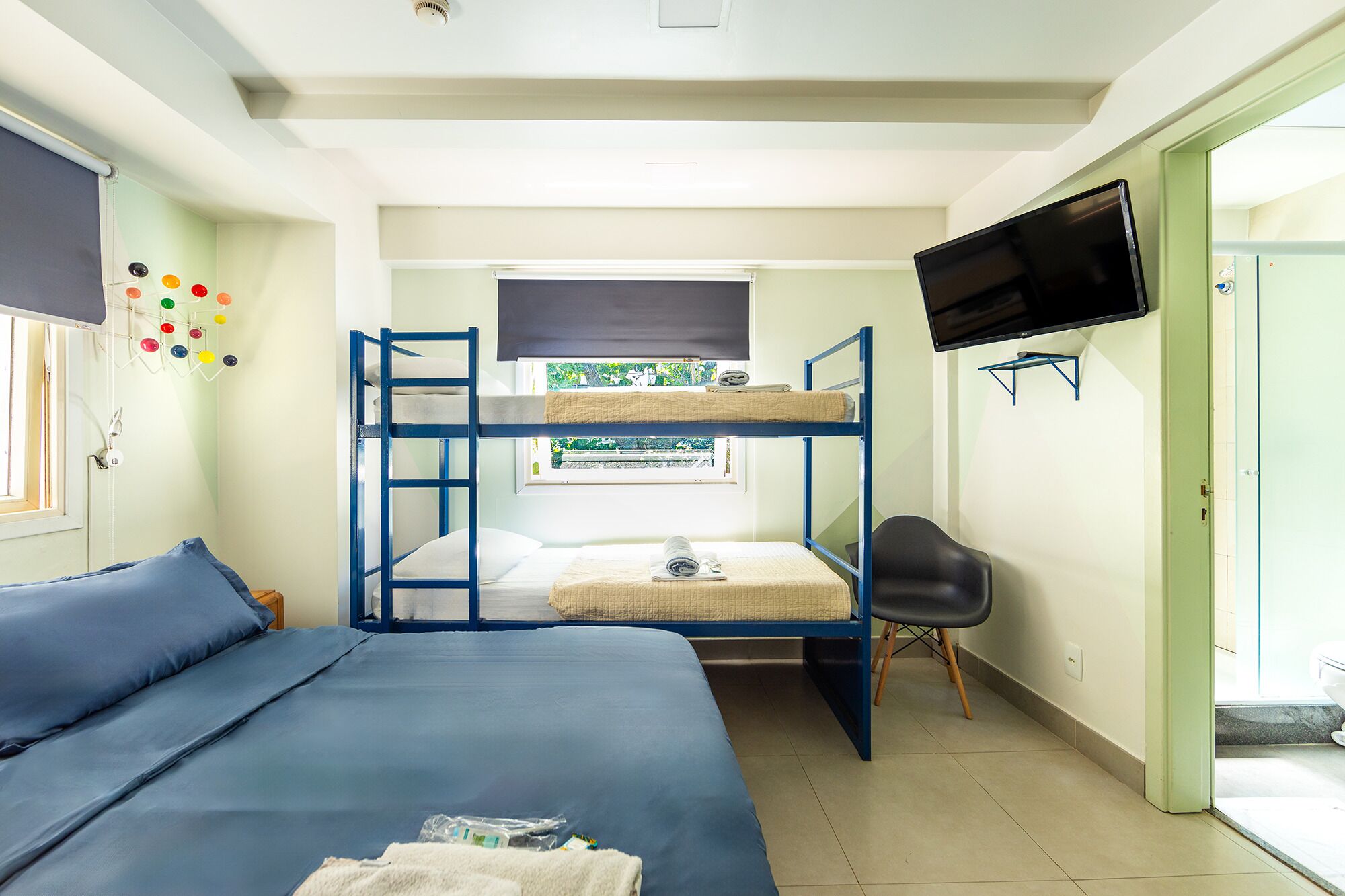 A dormitory room with bunk beds, a TV, a chair, and a window with a view of greenery.