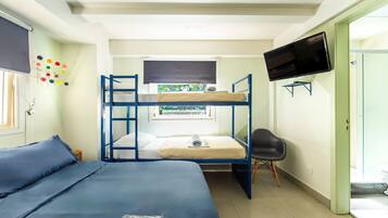 A dormitory room with bunk beds, a TV, a chair, and a window with a view of greenery.