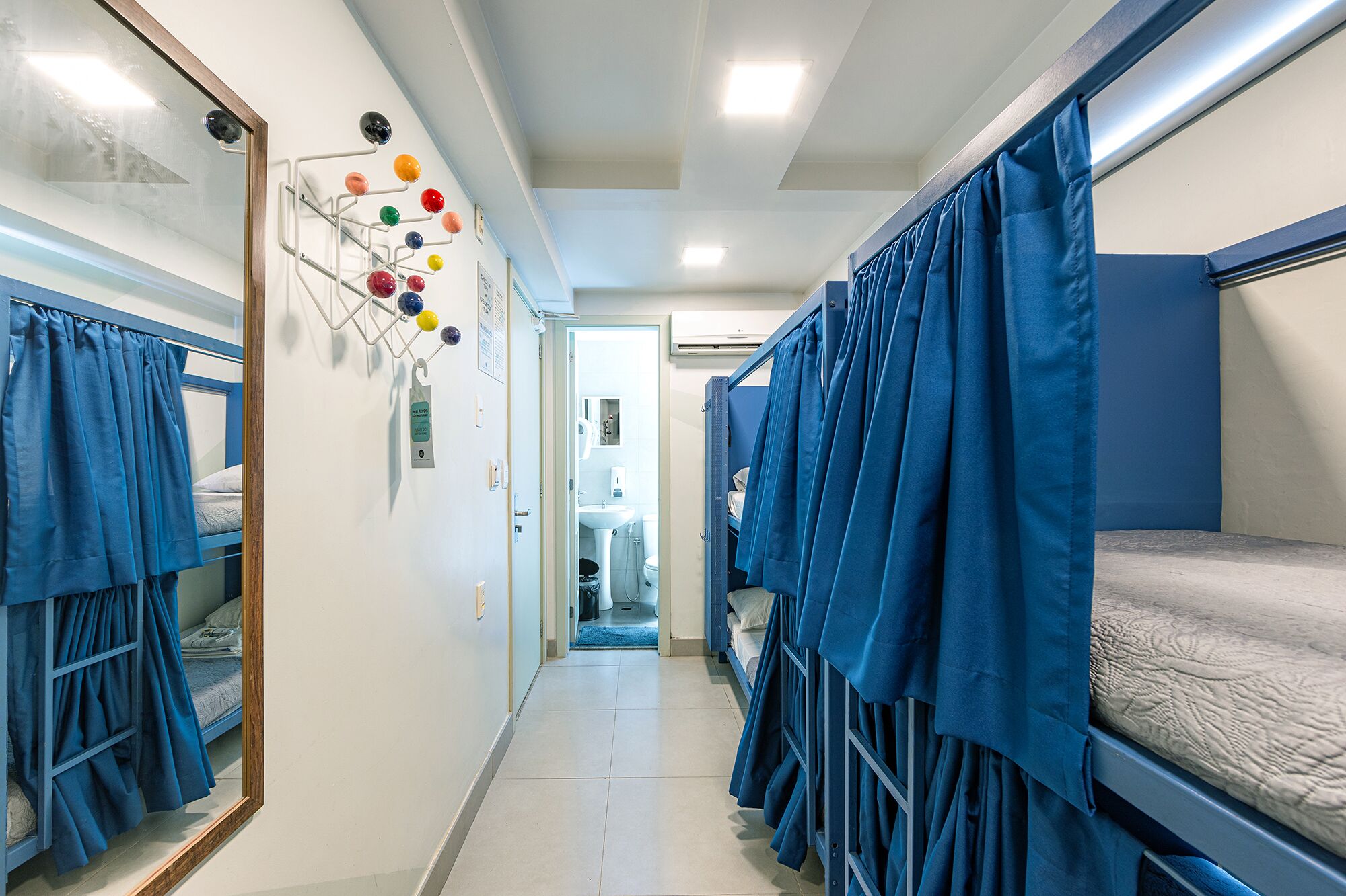 Un pasillo estrecho con literas a ambos lados, un tendedero de pared con bolas de colores, y un baño visible con lavamanos e inodoro.
