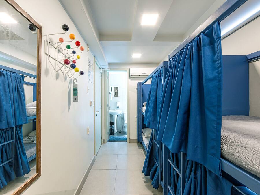 Un pasillo estrecho con literas a ambos lados, un tendedero de pared con bolas de colores, y un baño visible con lavamanos e inodoro.