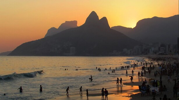 Una playa con gente disfrutando del atardecer y las olas, con montañas al fondo.