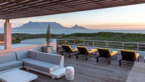 Eine Holzterrasse mit einer Couch und Liegestühlen, mit Blick auf eine Küstenlandschaft mit Bergen.