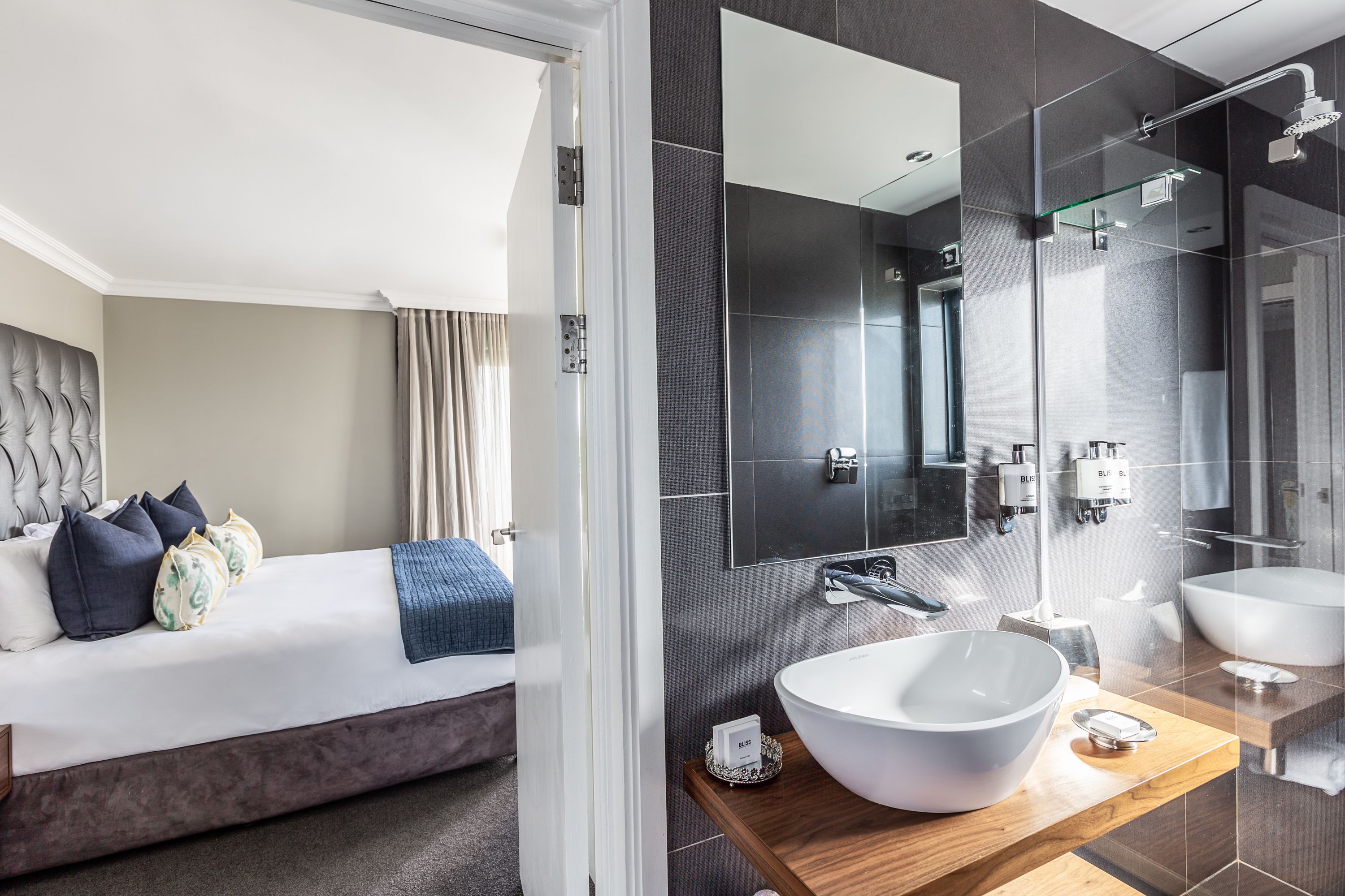 A modern hotel room with a large bed, a wooden vanity with two sinks, and a large mirror.
