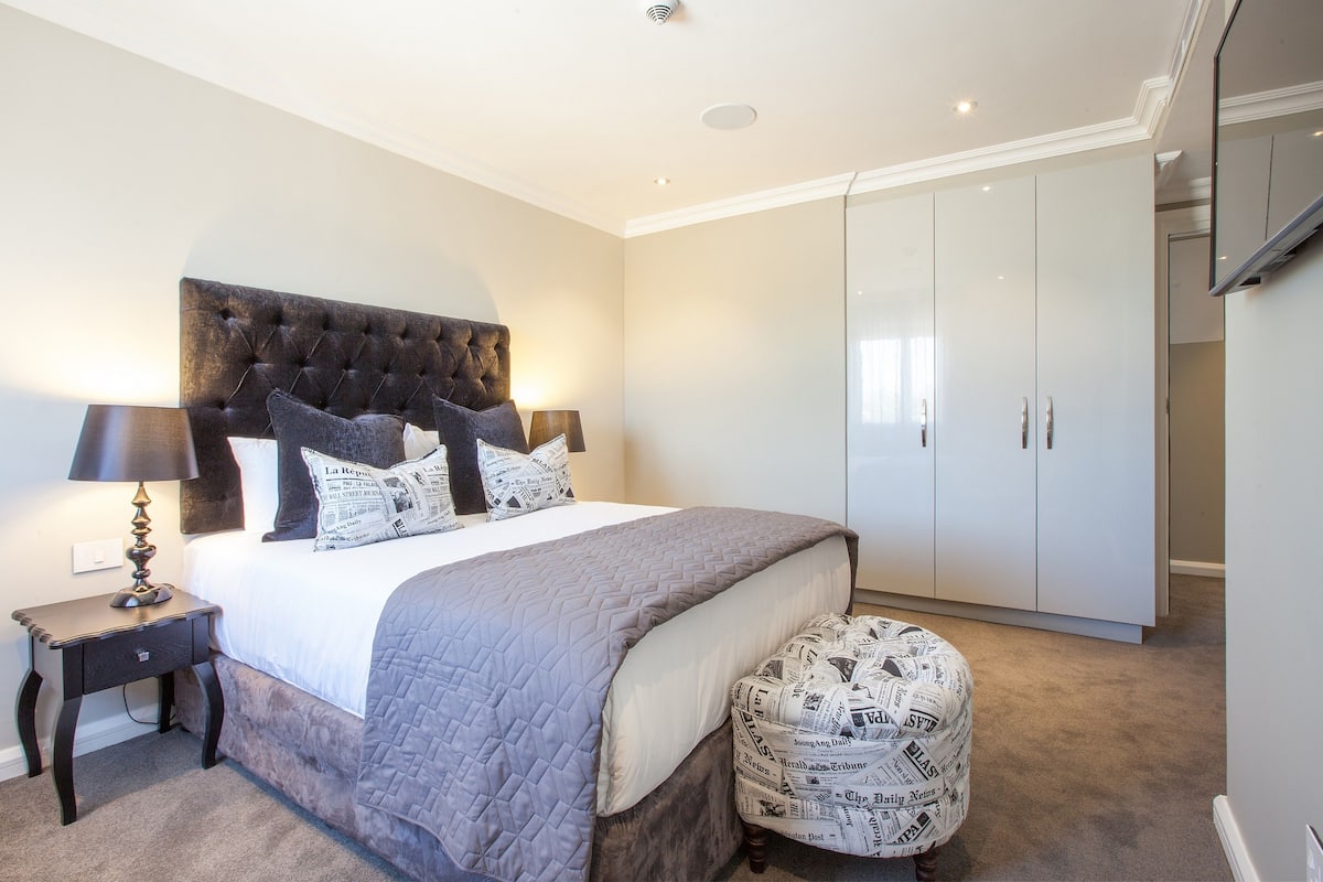 Une chambre avec un grand lit, une table de chevet, une armoire et un pouf à motifs.