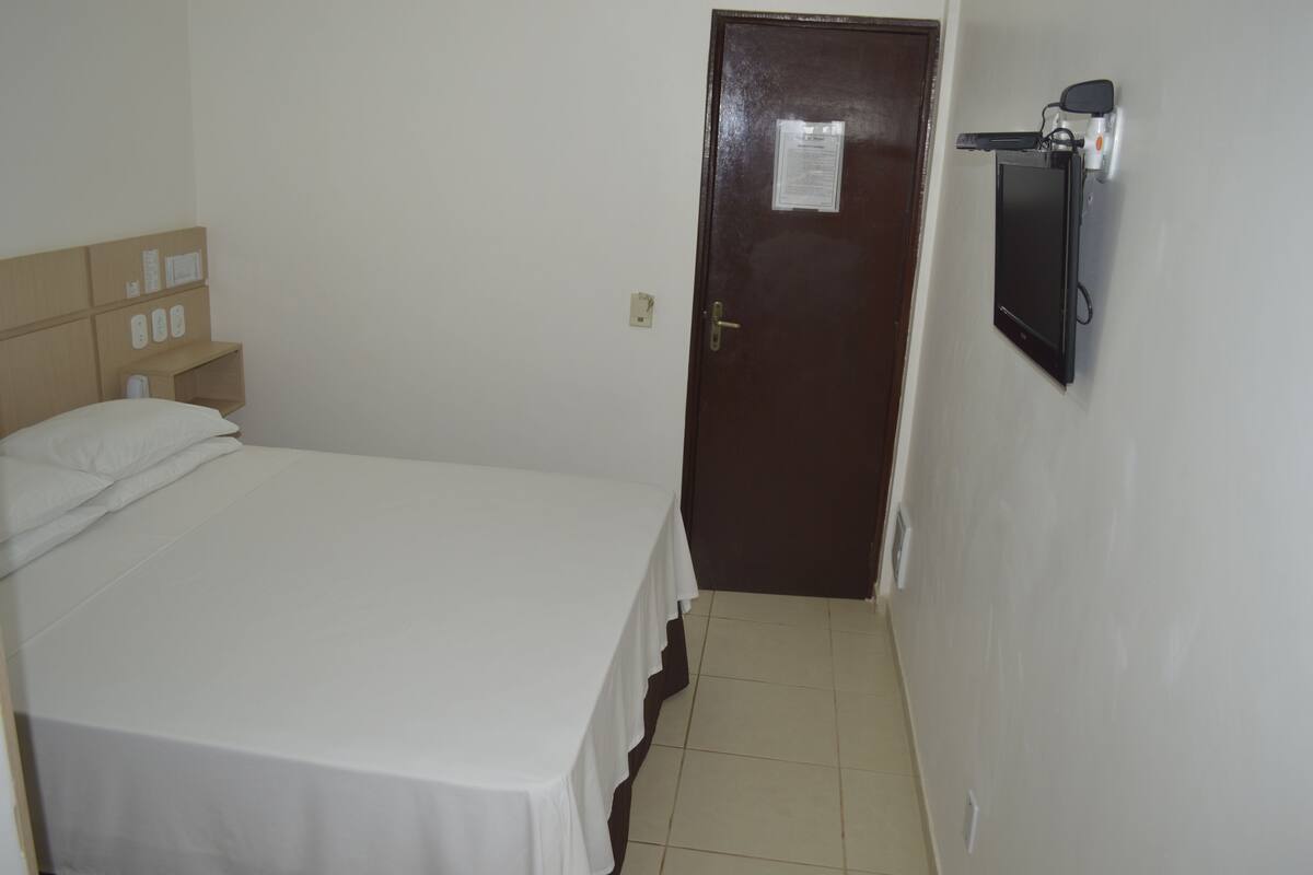 A small, clean room with a single bed, a wooden headboard, a door, and a mounted TV.