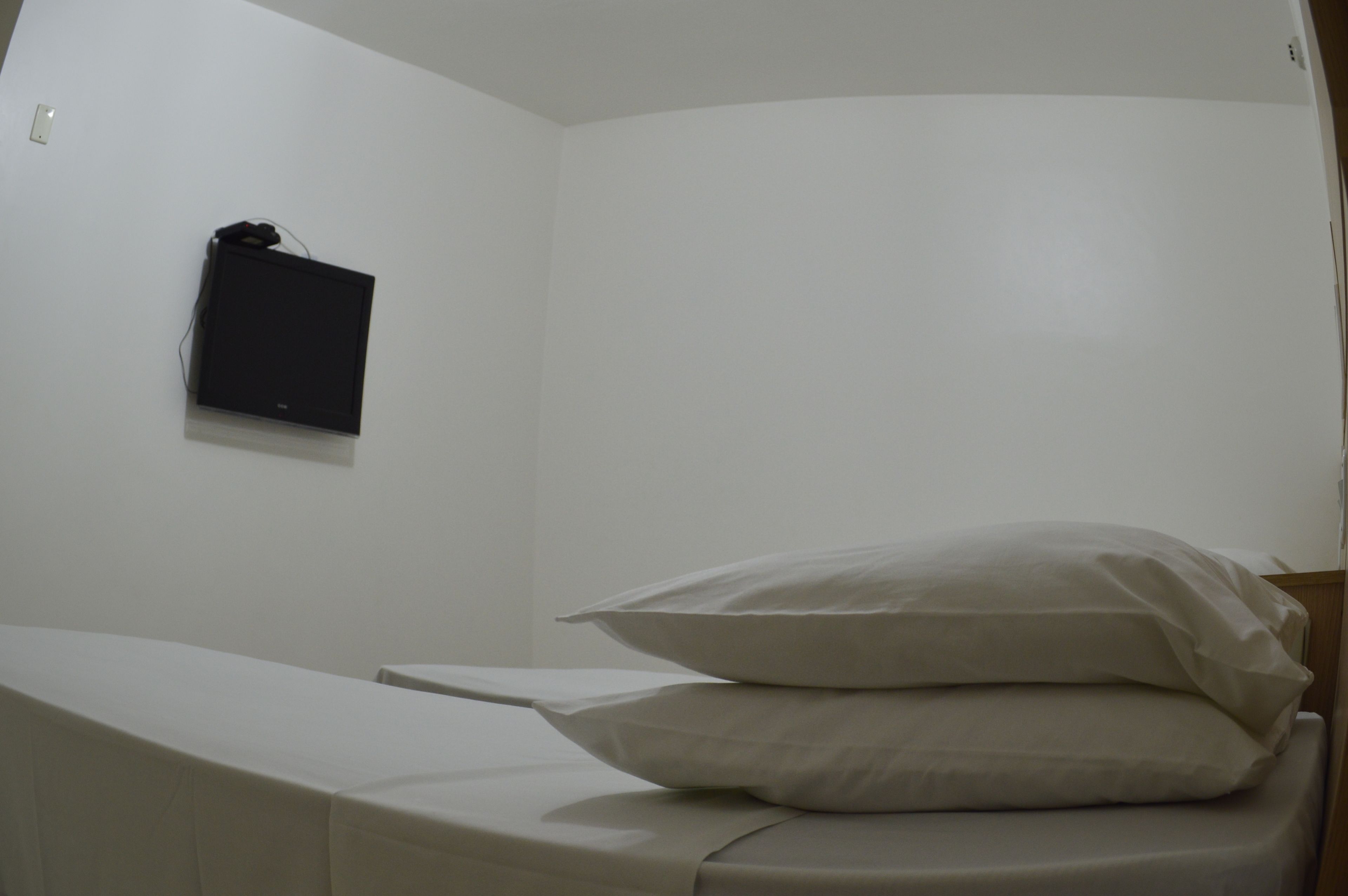 A small, single-bed room with a white wall, a flat-screen TV mounted on the wall, and a neatly made bed with white bedding.