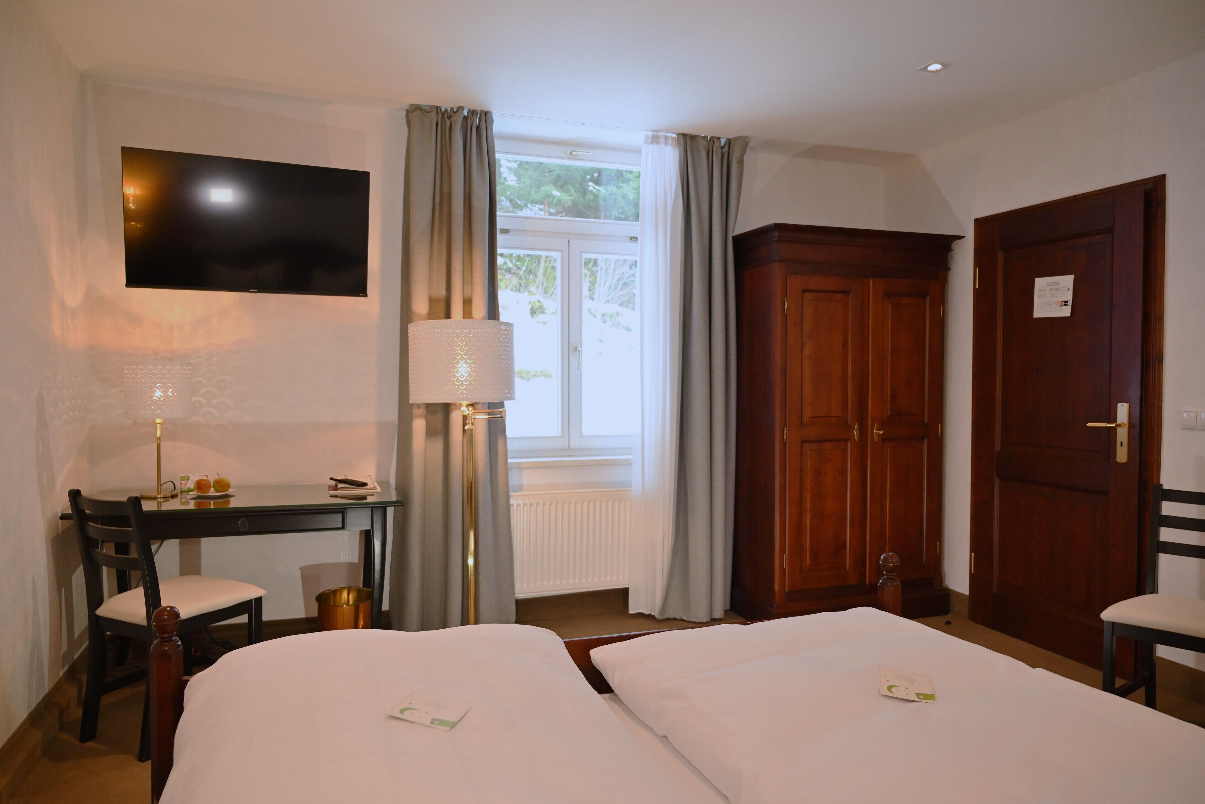 Habitación de hotel con dos camas, un escritorio con silla, un televisor montado en la pared, una ventana con cortinas y un armario de madera.